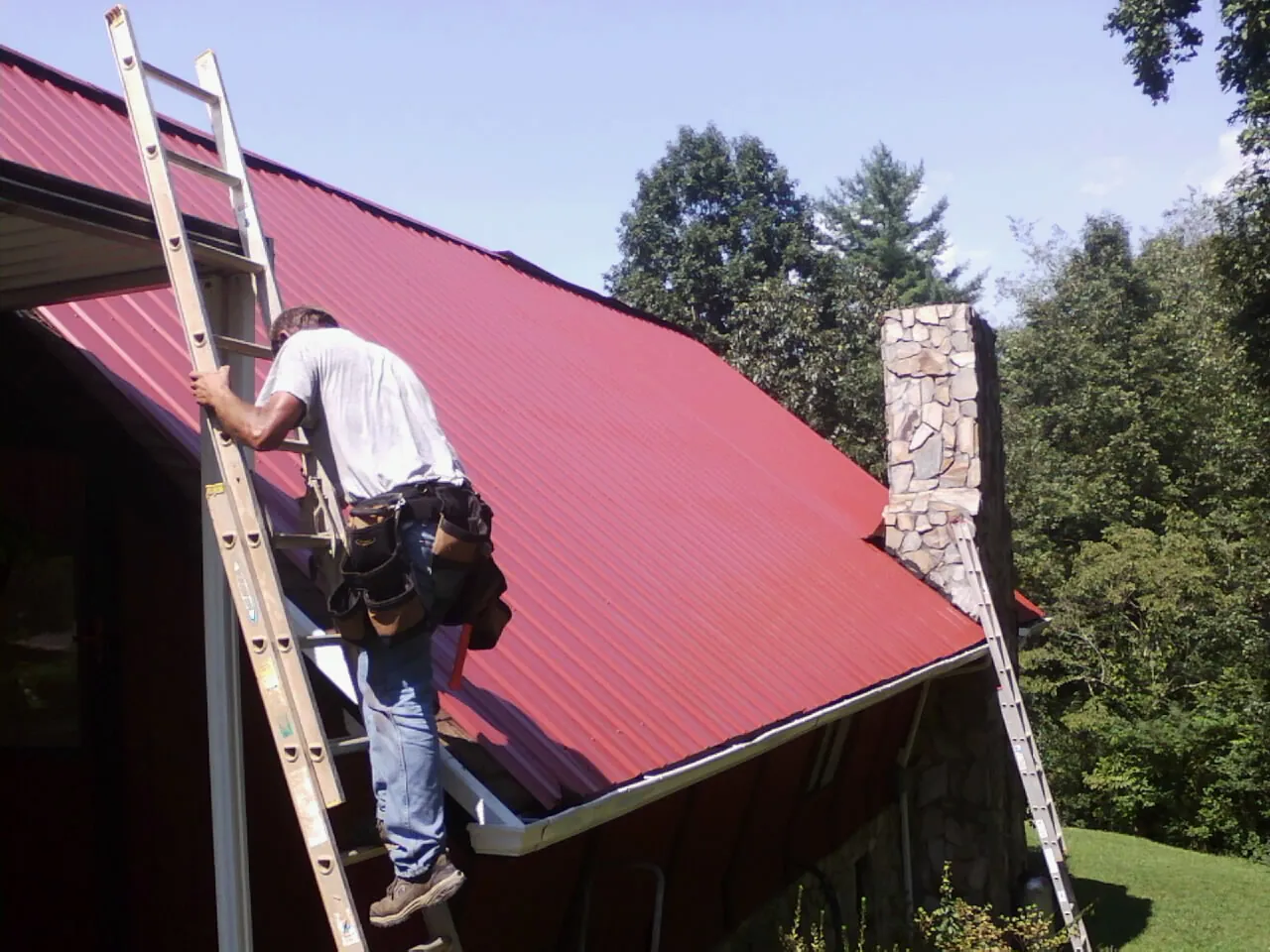 Roofer on a ladder beside a red metal roof and stone chimney — Soffit & Fascia Repair by Roofing Brothers in Republic